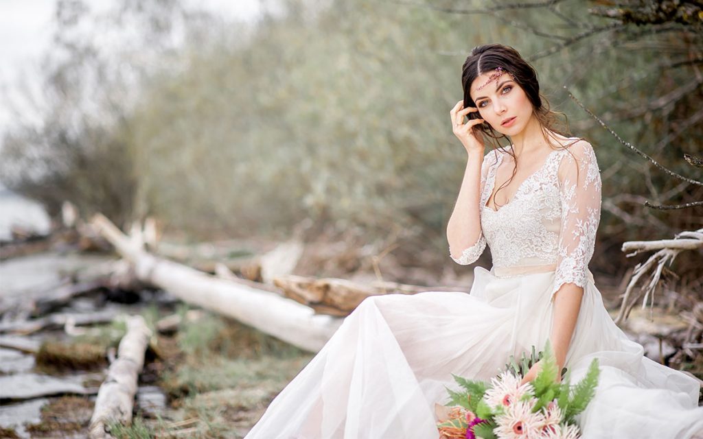 Braut sitzt mit Brautstrauß auf Treibholz am Seeufer